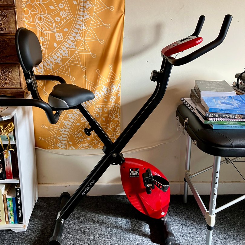 Red folding exercise bike with a supportive seat and backrest, positioned beside spiritual books and an orange mandala wall hanging.