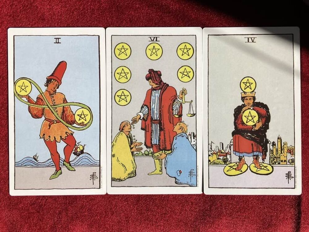 Three Rider-Waite Tarot cards—the 2, 6 and 4 of Pentacles—arranged on red fabric.