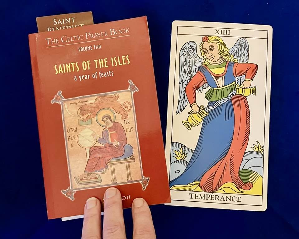 Saints of the Isles prayer book beside a Tarot de Marseille Temperance card depicting an angel pouring between two vessels, on blue cloth.
